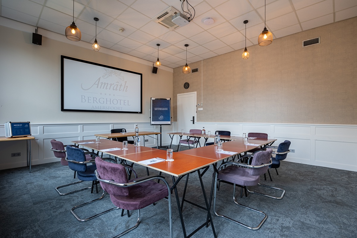 Vergaderzaal in Amrâth Berghotel Amersfoort opgesteld in een u-vorm met houten tafels met flessen water en grijze stoelen. Voorin de ruimte staat een centrale tafel met daarachter een beamer en een flipover 