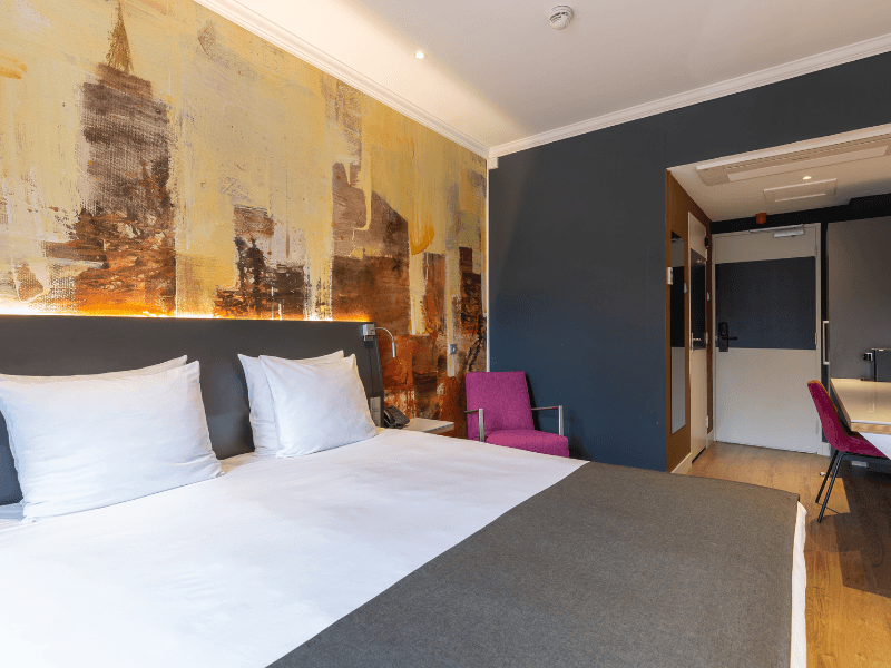 Comfort double kamer in Amrâth Berghotel Amersfoort met twee eenpersoonsbedden met een grijs sprei en een gekleurde muur erachter met daarnaast een paarse fauteuil