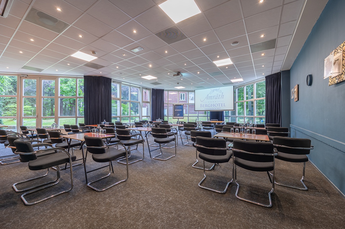 Vergaderzaal Amrâth Berghotel Amersfoort met houten tafels met daaromheen vier zwarte stoelen voor in de ruimte staat een centrale tafel met een beamer en een flipover naast een blauwe muur