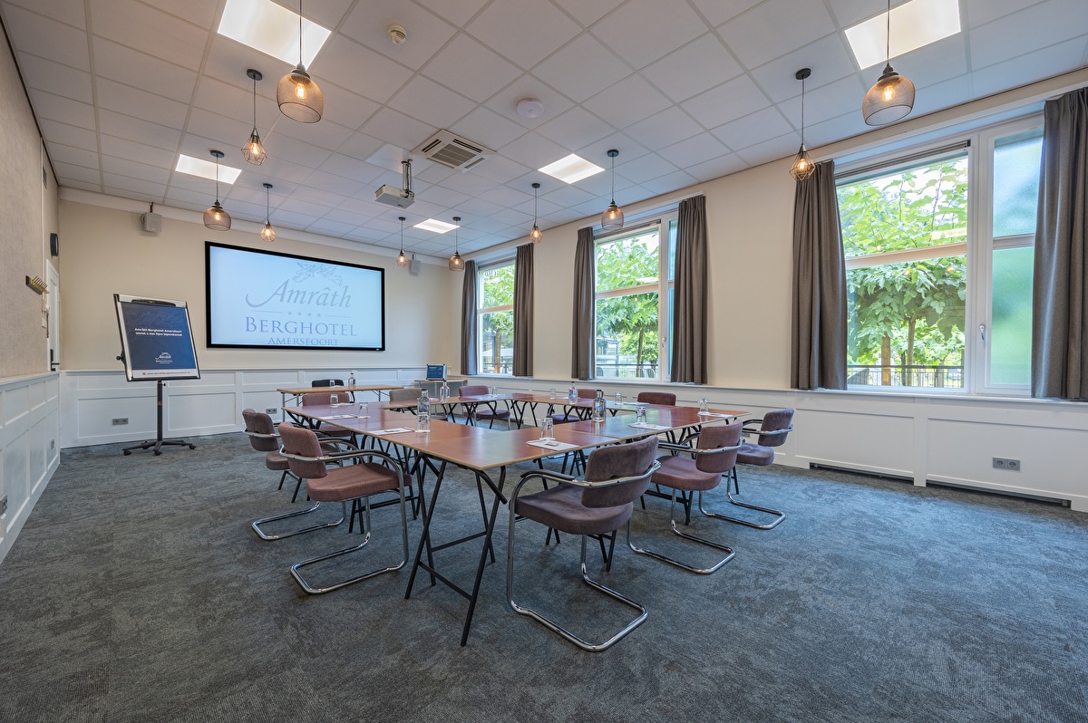 Vergaderzaal Amrâth Berghotel Amersfoort met houten tafels en paarse stoelen opgesteld in een vierkant. Voor in de ruimte staat een centrale tafel met daarachter een beamer en een flipover