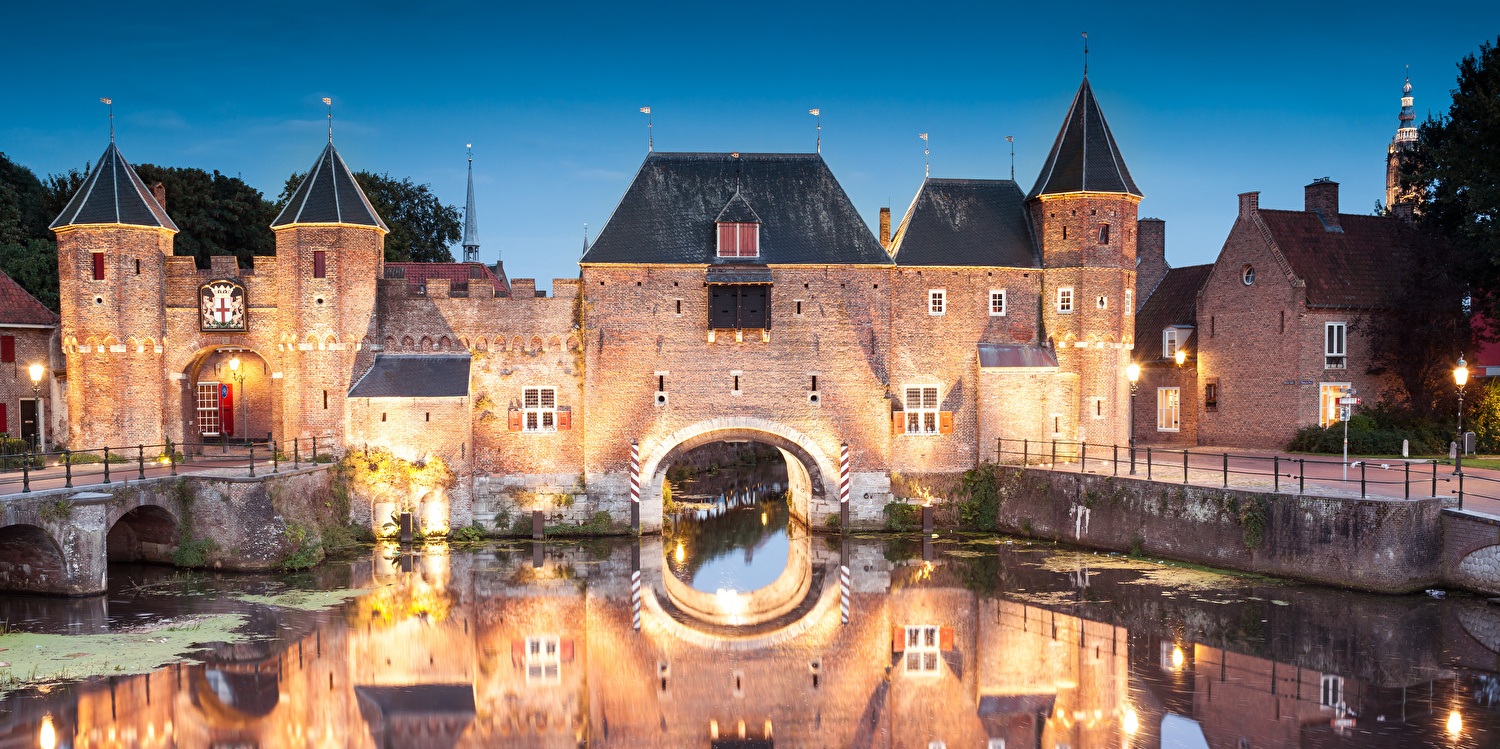 De middeleeuwse Koppelpoort in Amersfoort bij avondlicht, prachtig weerspiegeld in het water van de stadsgracht, met warme verlichting die de bakstenen poort en torens sfeervol accentueert.