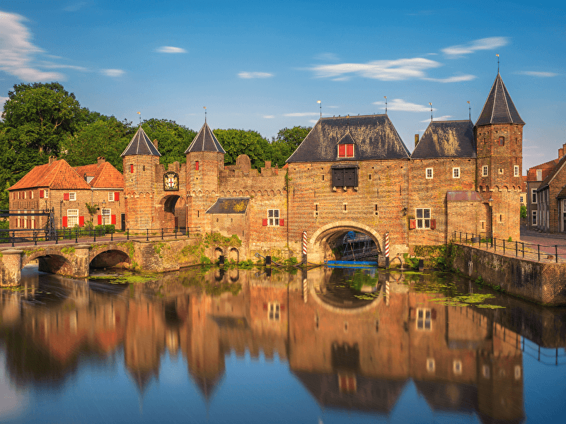 Koppelpoort Amersfoort omringd met een brug over water, met groene bomen en een blauwe lucht.
