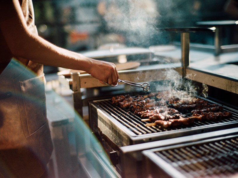 Persoon grilt vlees op een professionele barbecue tijdens een zomerse BBQ-avond op het terras van Amrâth Berghotel Amersfoort, met rook en zonlicht voor een sfeervolle uitstraling.          Vraag ChatGPT