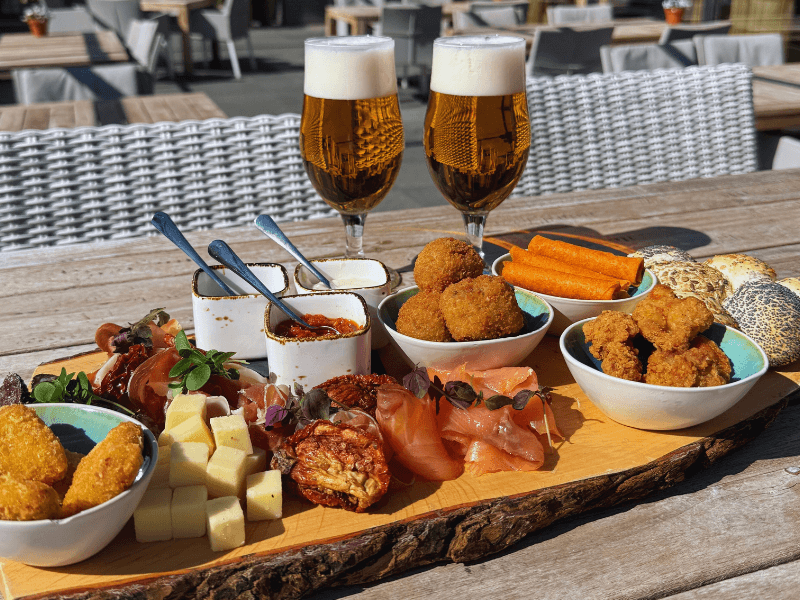 Plateau Amrâth geserveerd op een houten plank met diverse luxe hapjes zoals kaasblokjes, charcuterie, bitterballen, kipstukjes, dips en broodjes, vergezeld van twee glazen bier op een zonnig terras.