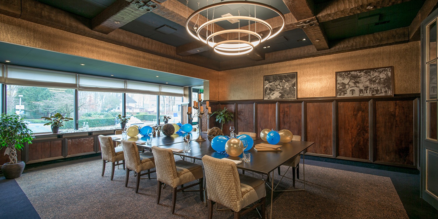 Meeting room in Amrâth Berghotel Amersfoort with a large wooden table with blue and gold balloons on it, beige chairs, with plenty of natural light.