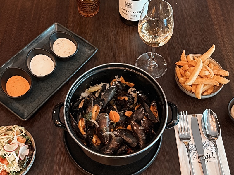 Geserveerde pan met verse mosselen in een zwarte pan, drie sausjes, friet, salade en een glas witte wijn (La Guirlande Sauvignon Blanc) op een donkerhouten tafel bij Amrâth Hotels.