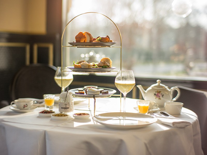 A set table with a high tea tiered stand, teapot, cups, and glasses, illuminated by sunlight