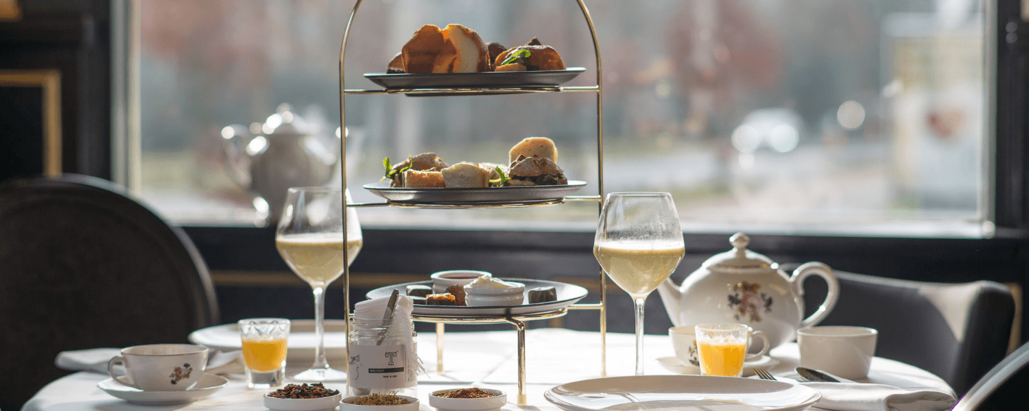 Een High Tea in Amrâth Berghotel Amersfoort opgedekt op een ronde tafel met een wit tafelkleed met daarop een etagiere met hartige en zoete hapjes met daarnaast servies een thee pot twee wijnglazen