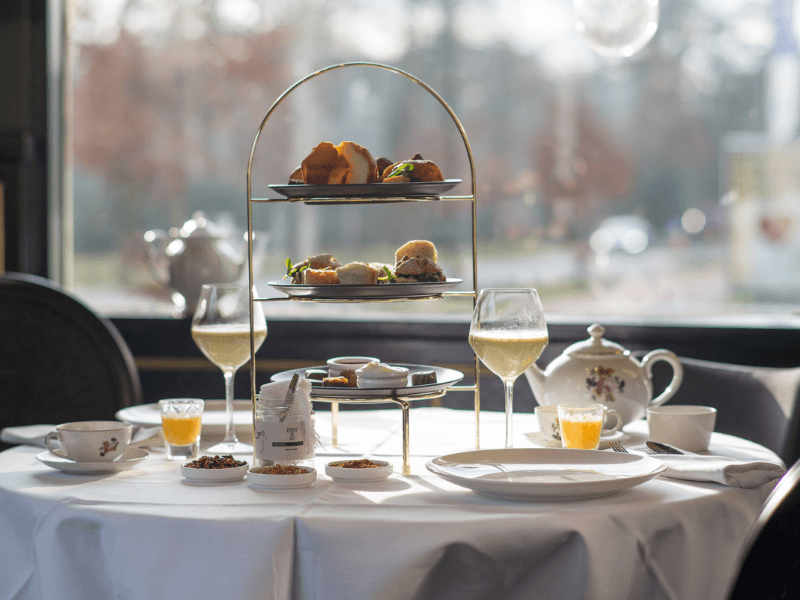 Ein High Tea im Amrâth Berghotel Amersfoort, gedeckt auf einem runden Tisch mit einem weißen Tischtuch, darauf eine Etagere mit herzhaften und süßen Häppchen, daneben Geschirr,