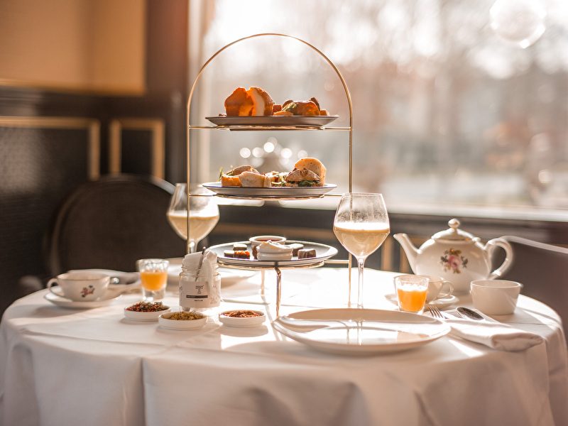 Ein High Tea im Amrâth Berghotel Amersfoort, gedeckt auf einem runden Tisch mit einem weißen Tischtuch, darauf eine Etagere mit herzhaften und süßen Häppchen, daneben Geschirr,