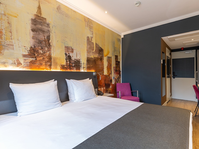 Comfort kamer in Amrâth Berghotel Amersfoort met een grijs sprei op bed, een roze stoel in de hoek en een geprint geel met zwart behang aan de muur