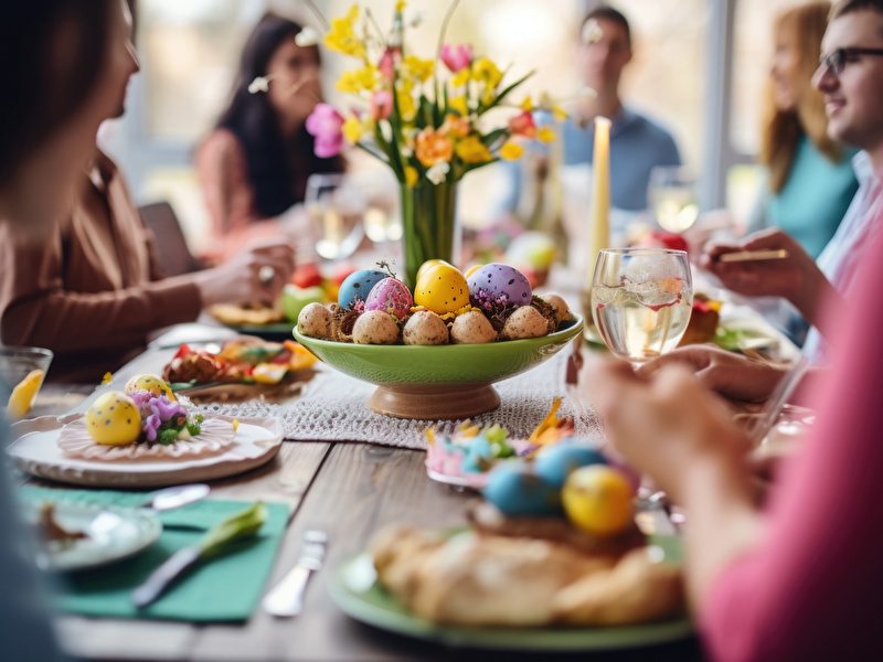 A group of people sits at a table surrounded by colorful Easter eggs, with delicious dishes on the table and a bunch of colored daffodils in the middle of the table.