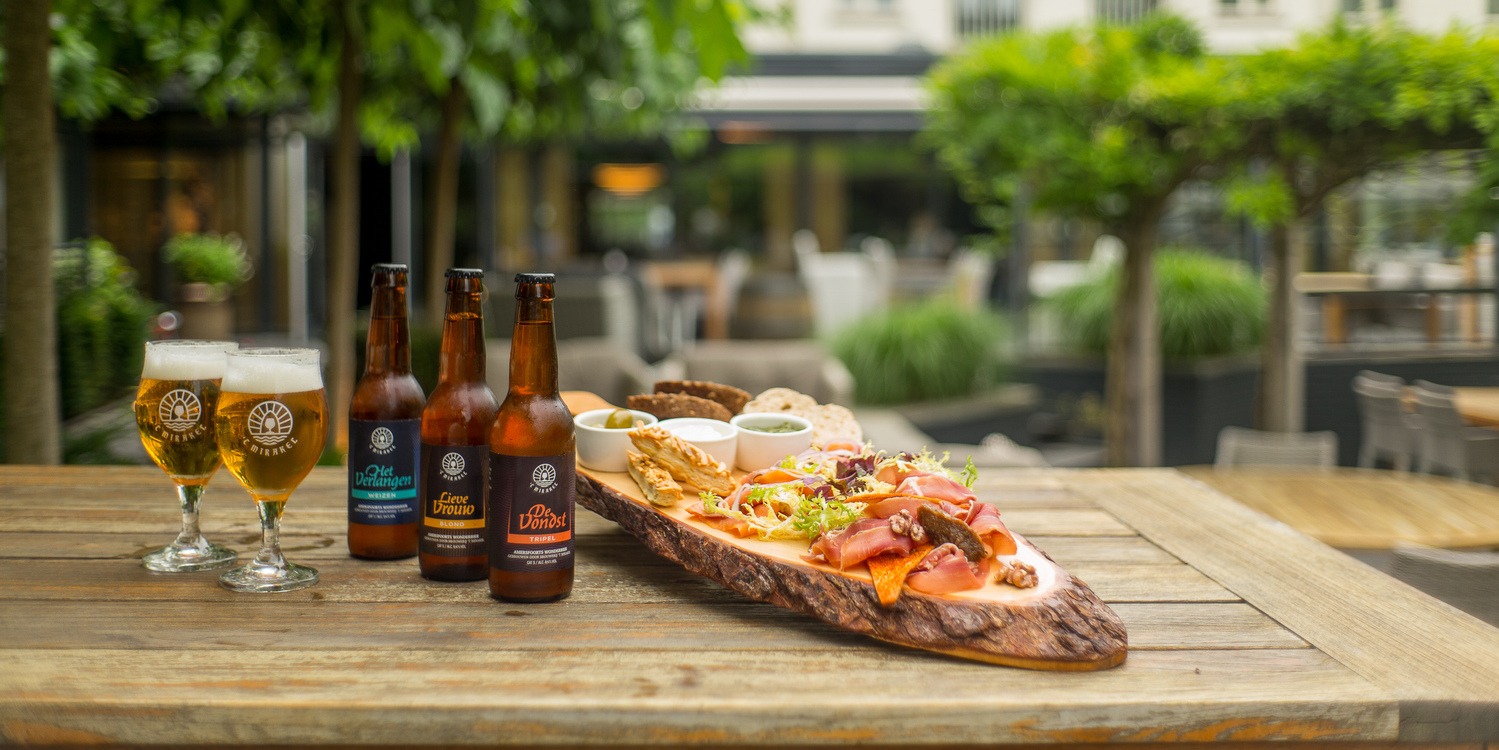 Plateau Amrâth served on a wooden board with various luxury snacks such as cheese cubes, charcuterie, bitterballen, chicken pieces, dips, and rolls, accompanied by two glasses of beer on a sunny terrace.