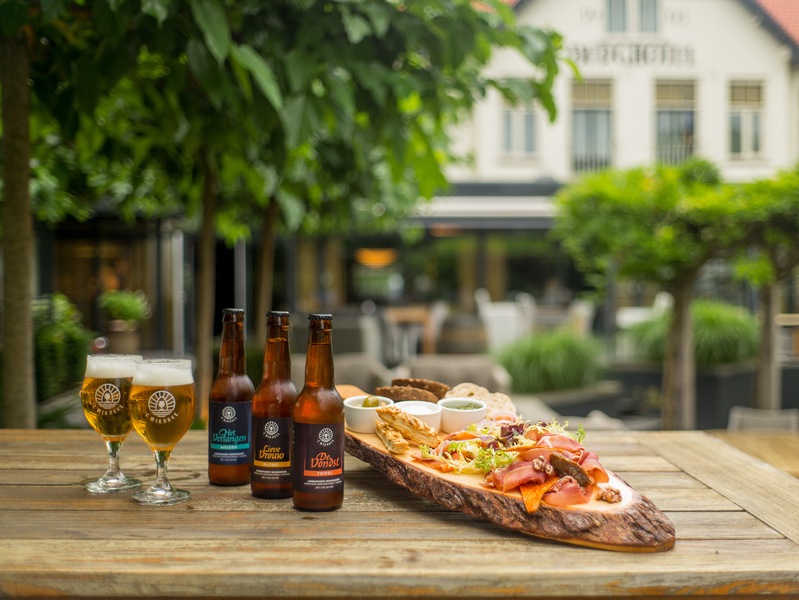 Amrâth platter served on a wooden board with various luxury snacks such as cheese cubes, charcuterie, bitterballen, chicken pieces, dips, and rolls, accompanied by two glasses of beer on a sunny terrace.