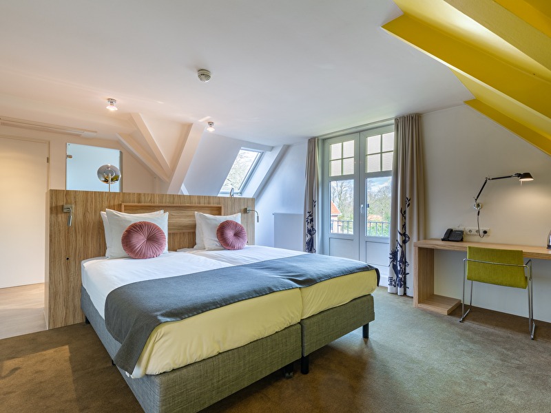 Superior room at Amrâth Berghotel Amersfoort with a large double bed with pink cushions, a desk with a chair, and a yellow wall.