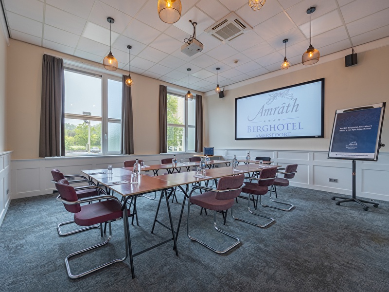 Meeting room in Amrâth Berghotel Amersfoort with a projector, tables and chairs arranged in a U-shape for a gathering.